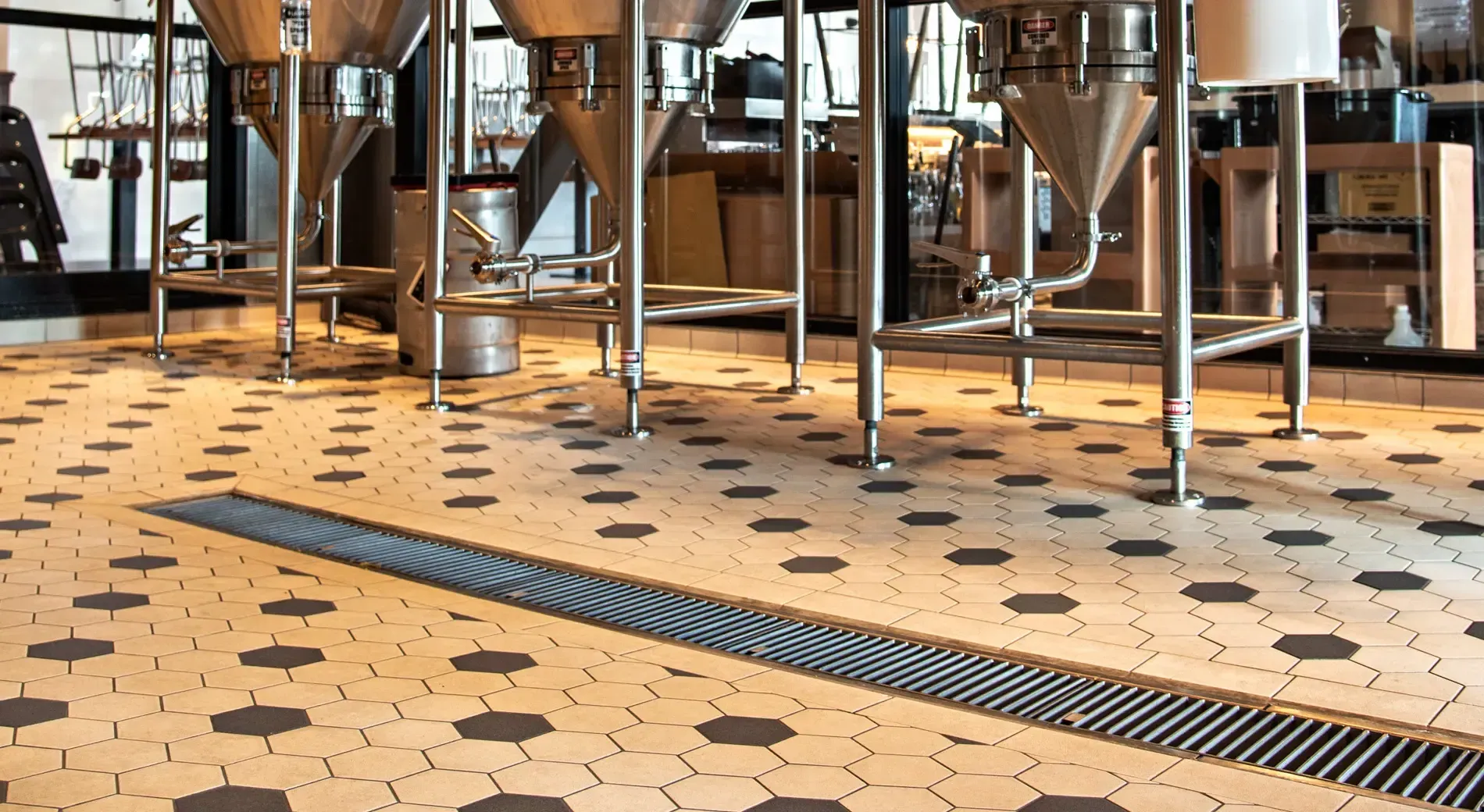 Stainless steel brewing tanks sit above a light-colored hexagonal tiled floor with a long, recessed drainage grate.