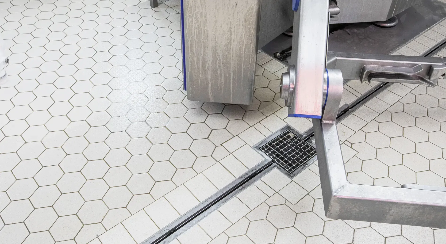 A close-up view of a white hexagonal tiled floor with a metal floor drain and industrial machinery in a clean facility.