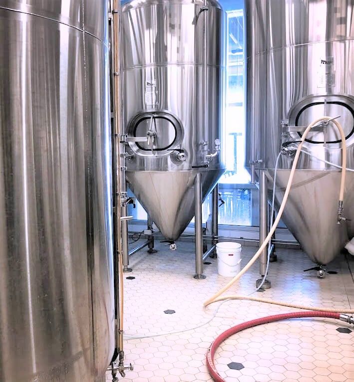 Three large, stainless-steel industrial fermentation tanks stand in a brewery on a tiled floor with hoses and a bucket.