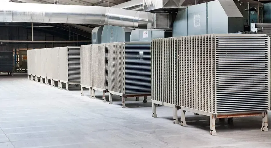 A row of industrial drying racks filled with trays sits on a factory floor under large ventilation ducts.
