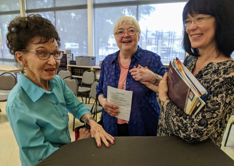 Three older women are standing around a table and smiling