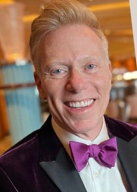 A man in a tuxedo and purple bow tie is smiling.