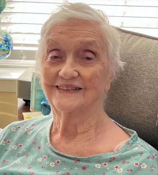 An elderly woman is smiling while sitting in a chair in front of a window.