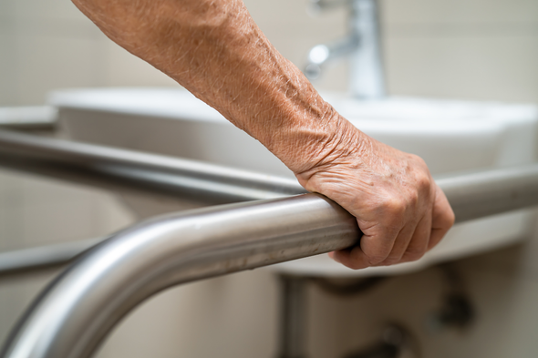 An elderly woman is holding onto a railing in a bathroom.