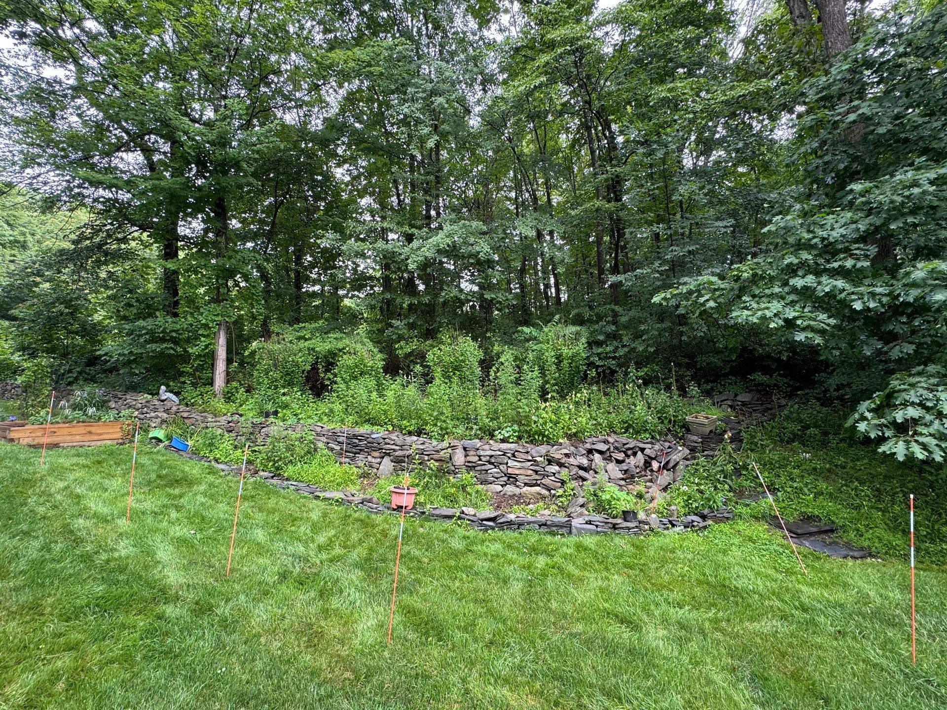 A stone-walled garden bed filled with plants, set against a backdrop of lush green trees, on a grassy lawn.