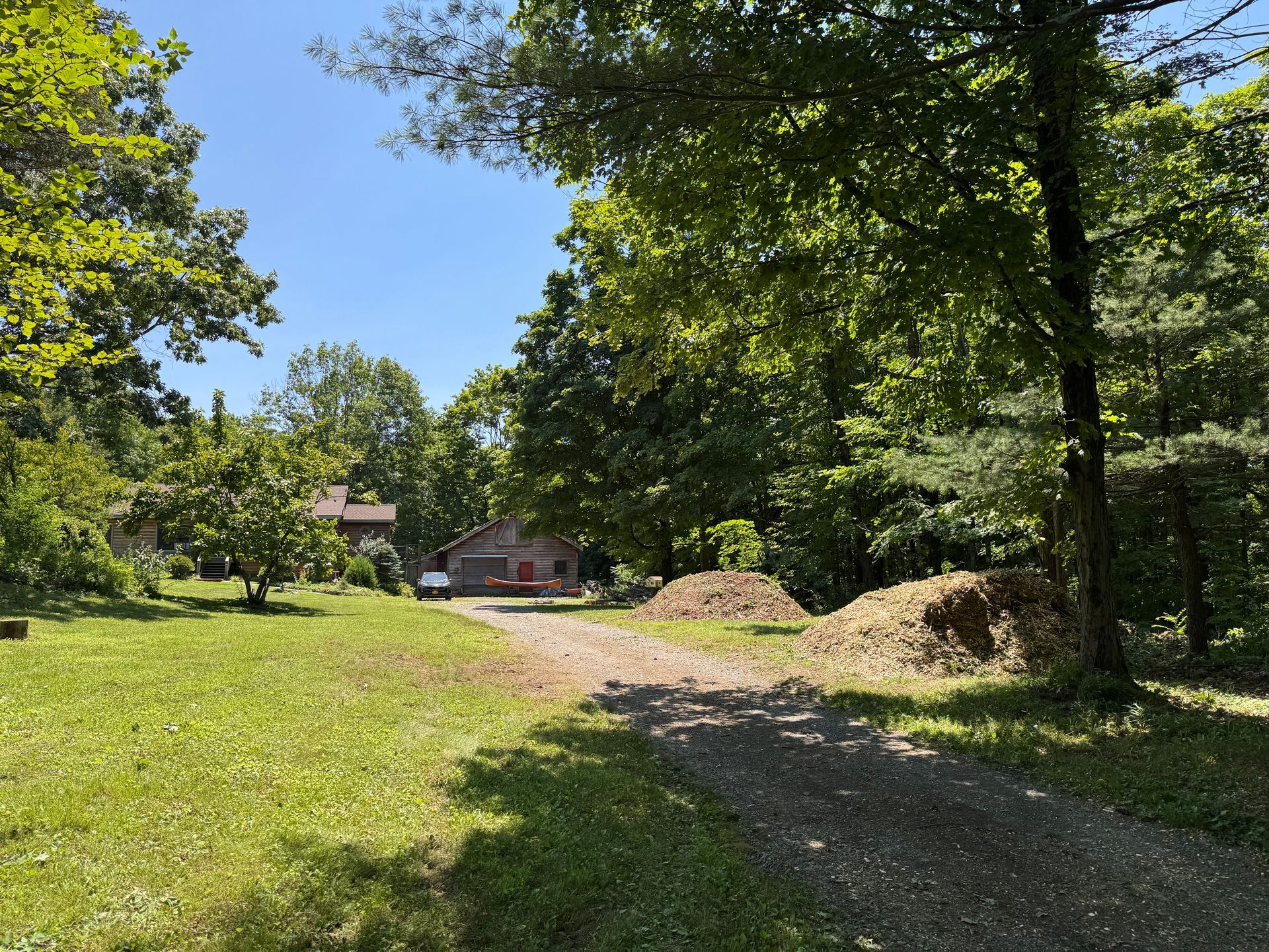 A gravel driveway leads towards two houses amidst a sunny, green landscape. Piles of wood sit to the right, and trees frame the scene.