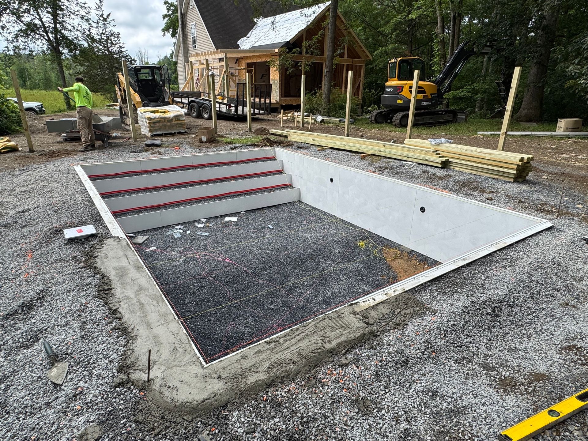 Construction site of a rectangular pool with built-in steps. Gray gravel base with white walls, a yellow leveler, and machinery present.