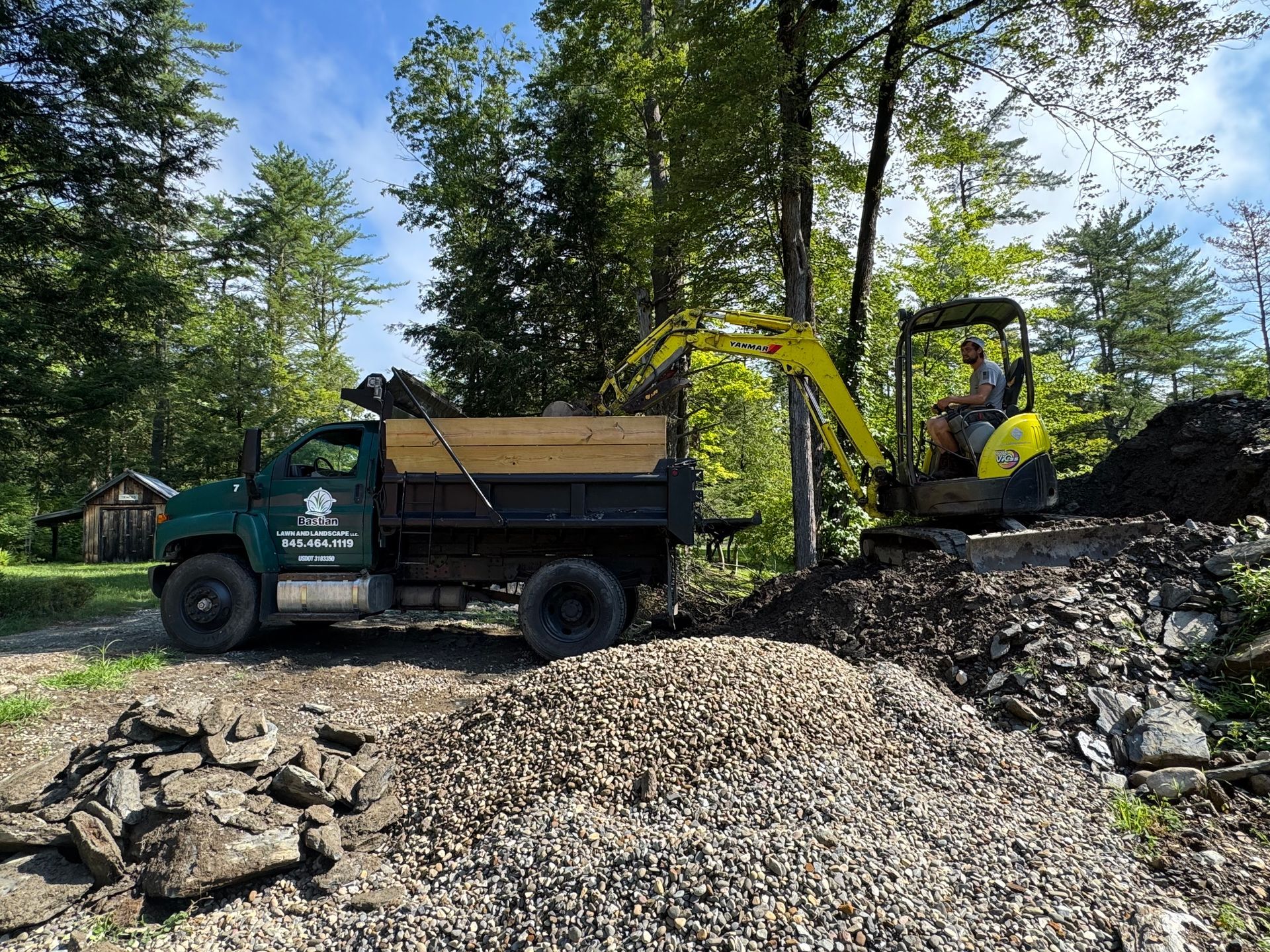 A mini-excavator loads gravel into a dump truck in a wooded area.  A person is in the excavator.