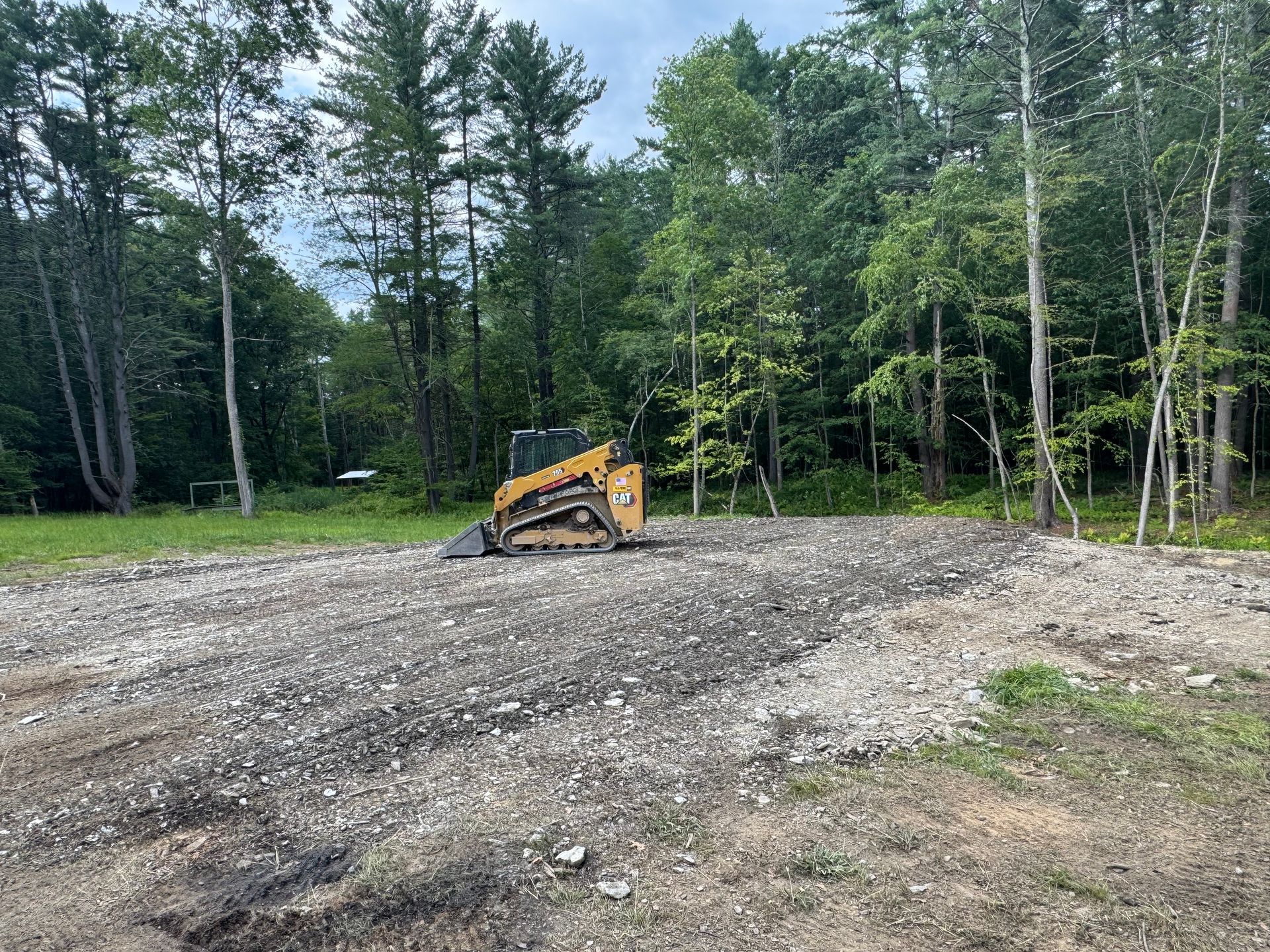 A skid steer sits on a gravel area clearing a space, with trees in the background.