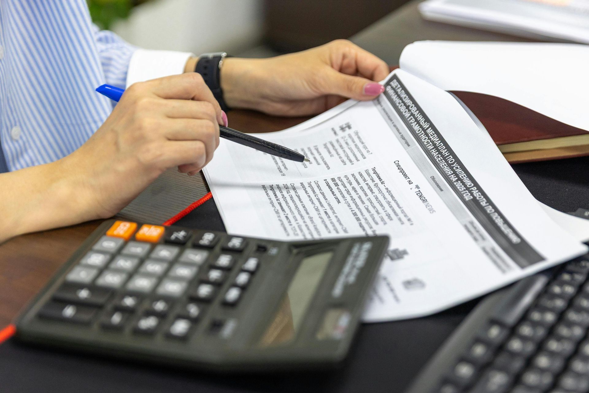 Hands at a desk use a pen to review a document next to a calculator and keyboard in an office setting.