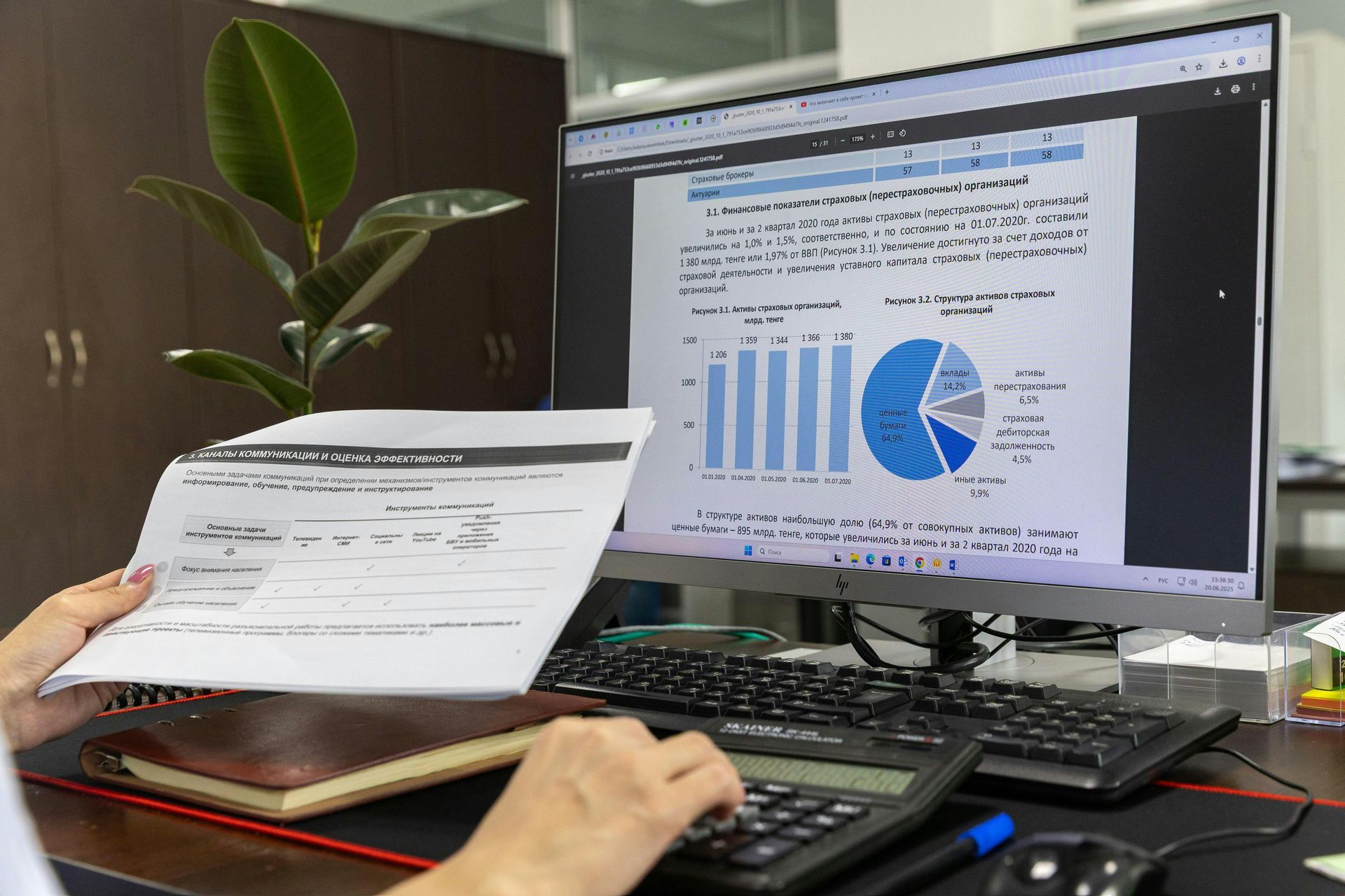 Hands using a calculator while holding a paper document, with a monitor displaying business charts in the background.