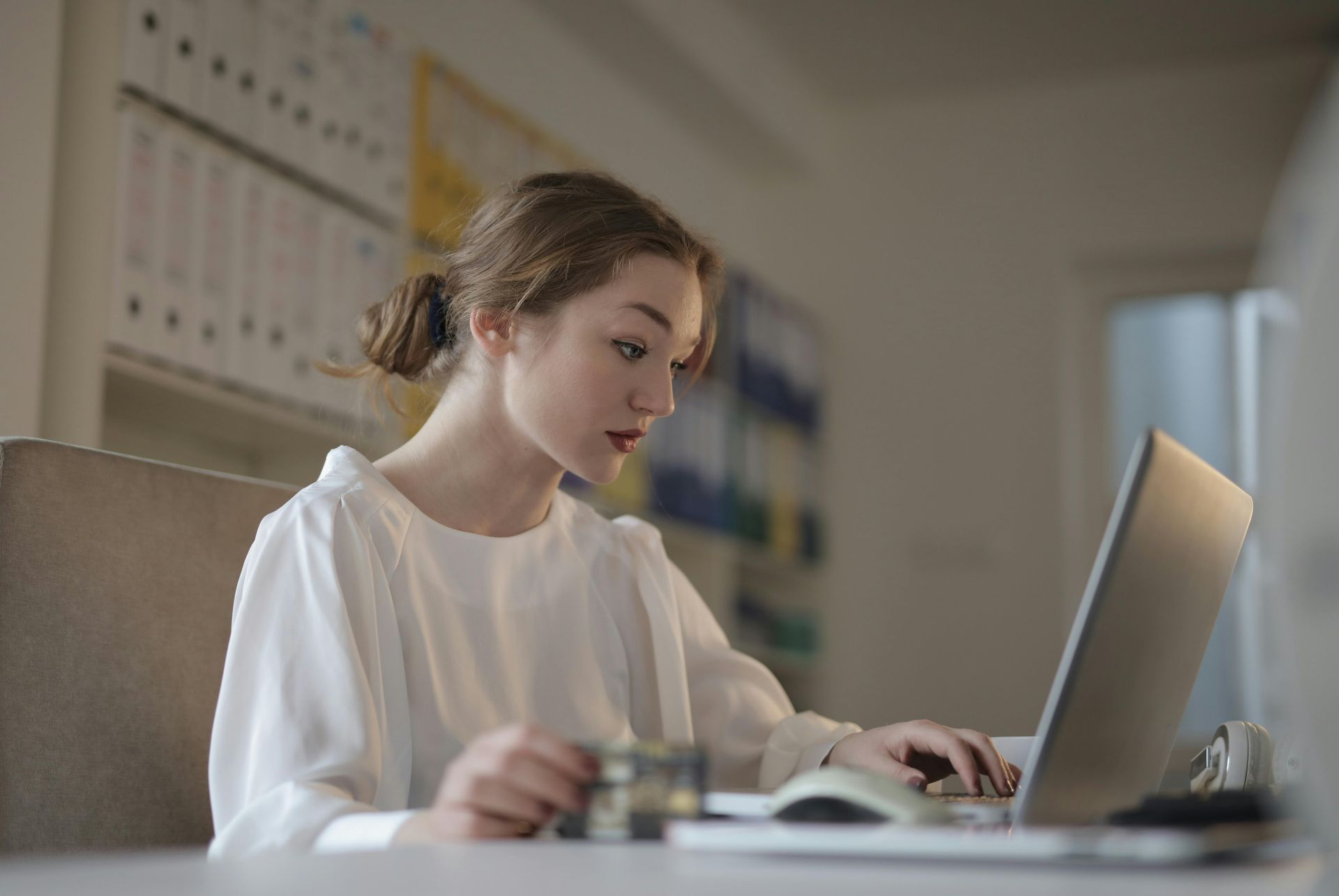 A person in a white top sits at a desk using a laptop in a brightly lit office with shelves of binders in the background.