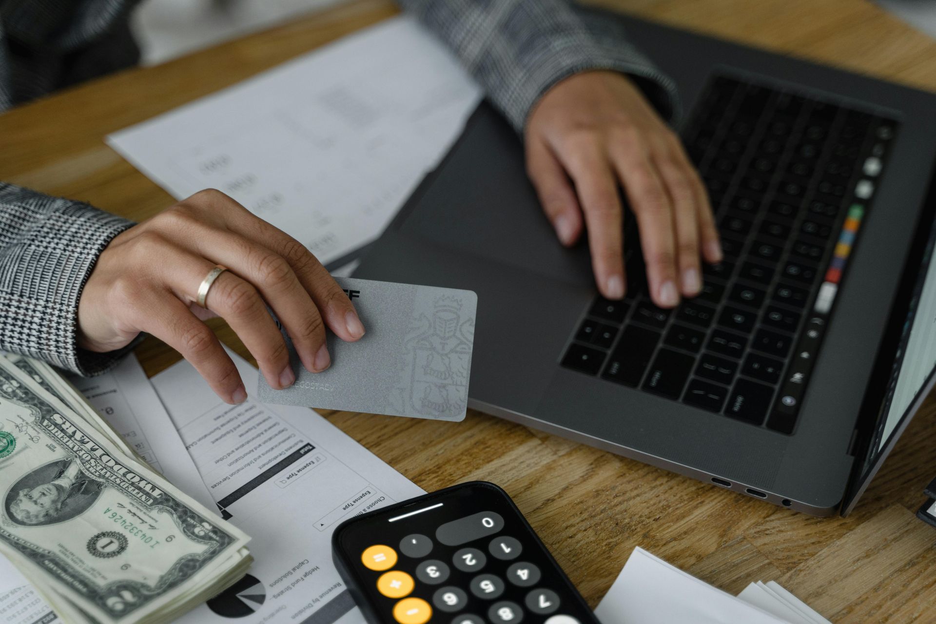A person holding a credit card while typing on a laptop next to a calculator and a stack of US dollar bills.
