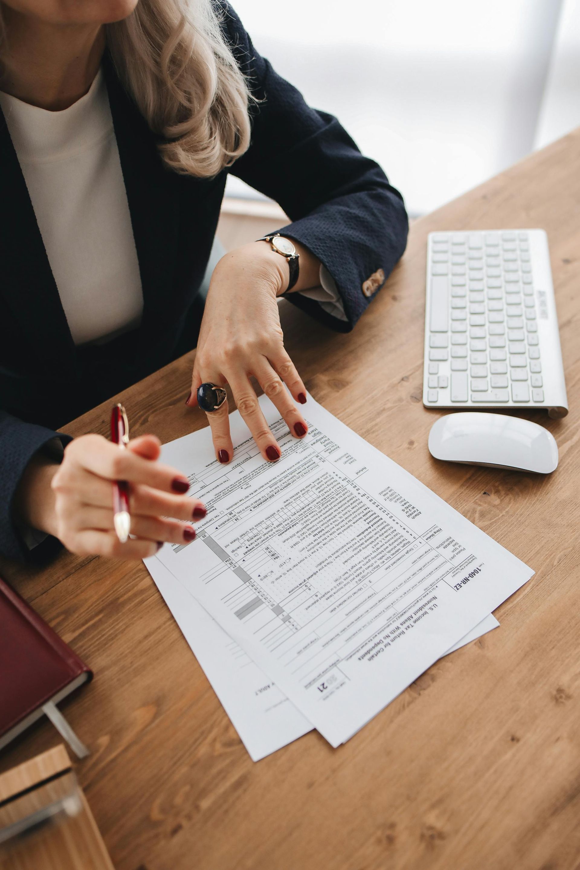 A person in a blazer sits at a wooden desk, reviewing a document with a pen in hand, next to a keyboard and mouse.