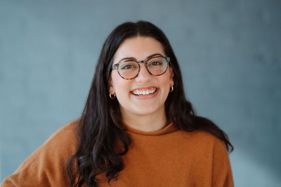 Alex Oswald wearing an orange sweater, glasses against a blue backdrop.