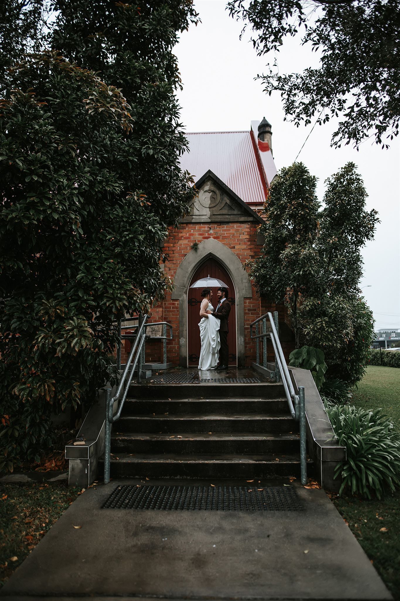 Bride and groom stand in front of an old church entrance in Northern NSW under an umbrella, on steps. Trees frame the shot.