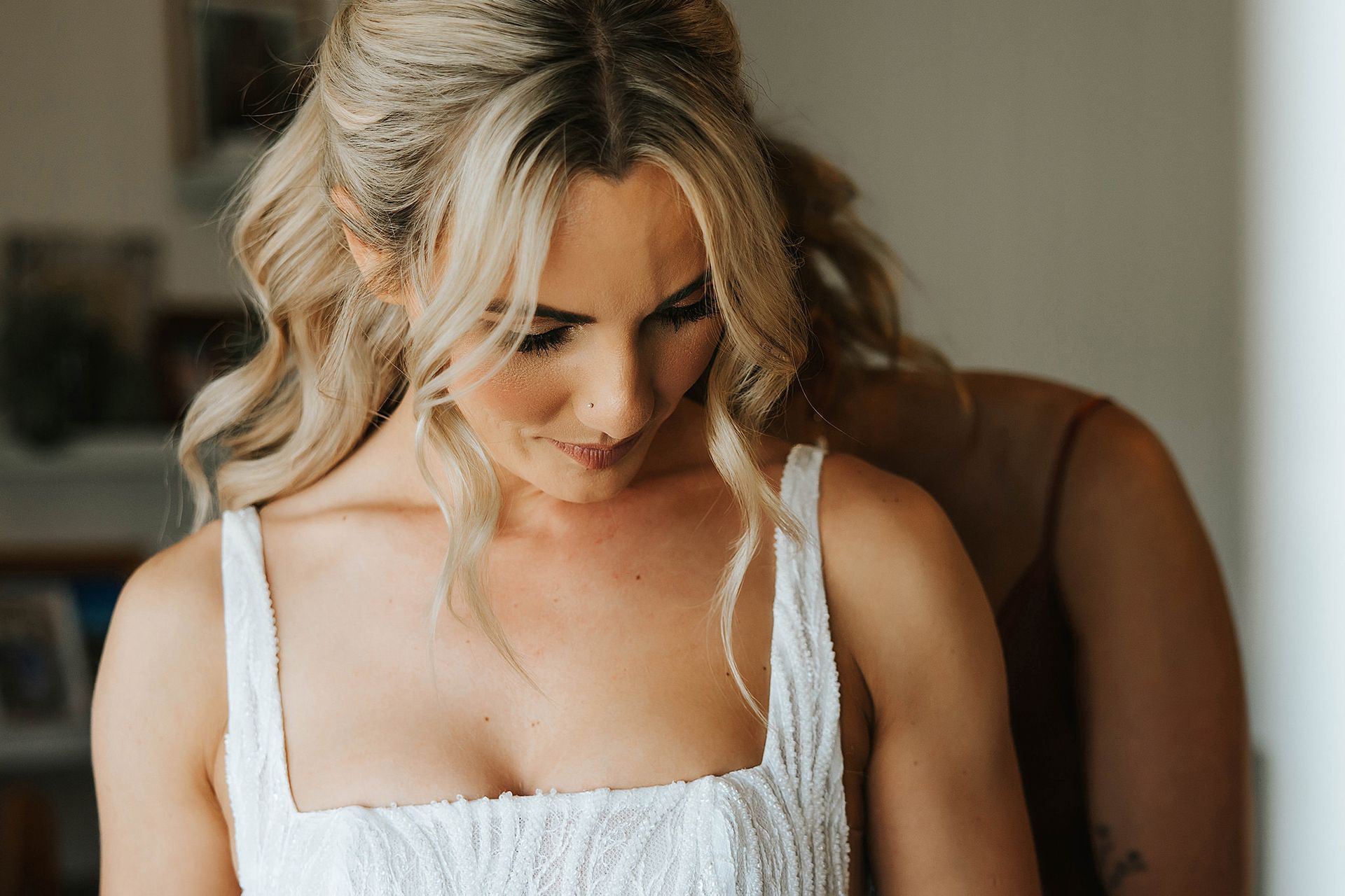 Blonde woman in a white dress, looking down with a bridesmaid adjusting her dress.