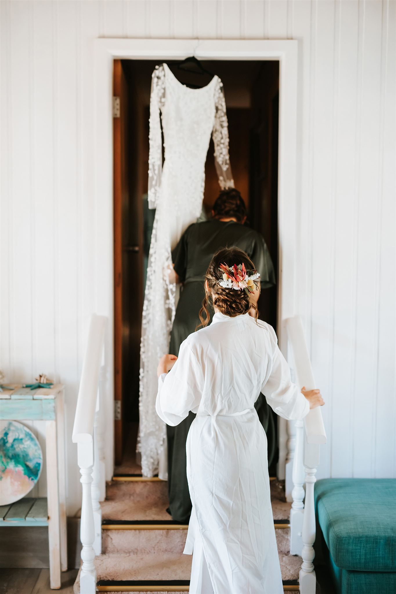 Bride in robe walks up stairs towards wedding dress hanging in doorway.