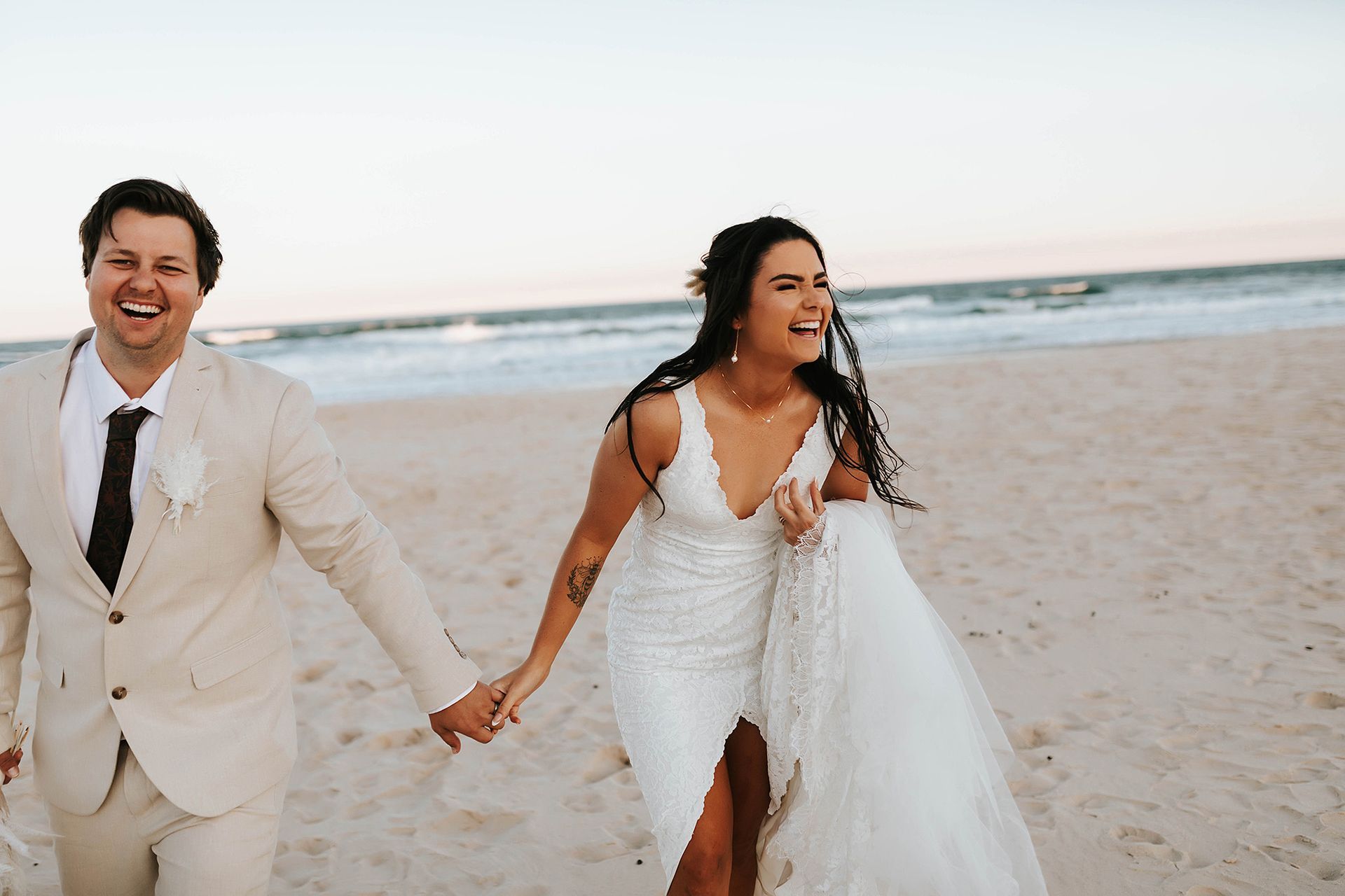Newlyweds laugh, holding hands, running on a sandy beach in Northern NSW.