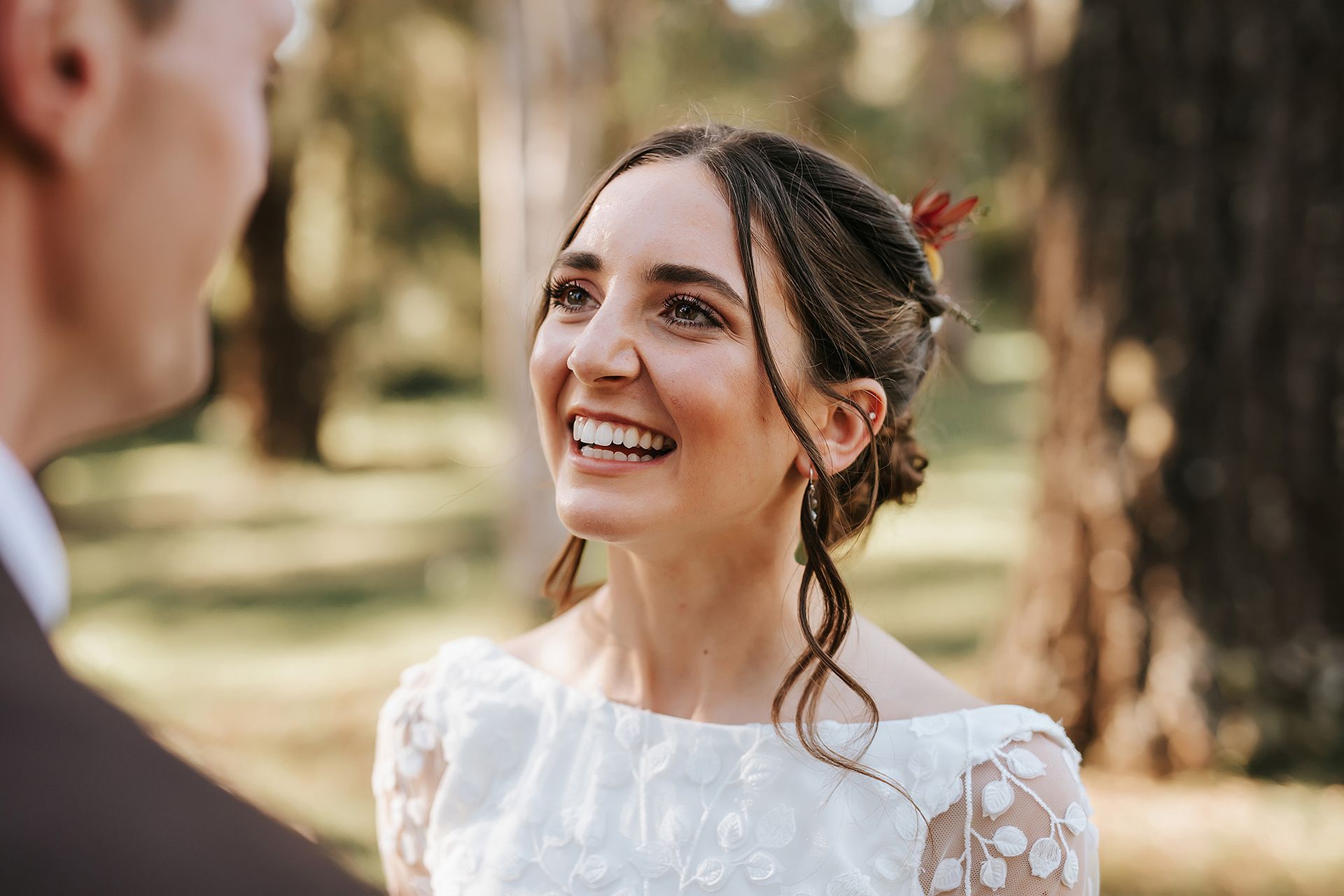 Bride smiles at groom, Northern NSW forest backdrop. She wears a white lace dress, updo with braids.