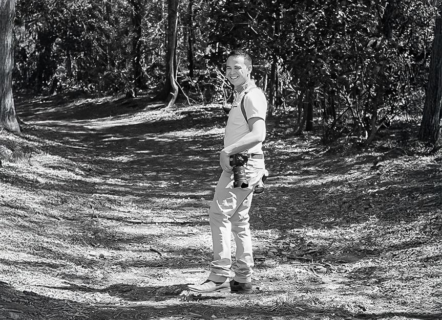 Denis Banks standing on a path in a forest, smiling, with a camera around their neck.