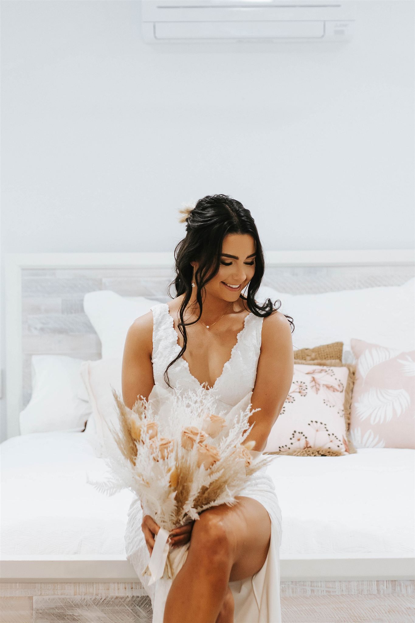 Woman in white wedding dress smiles, holding bouquet, sitting on bed with pillows.