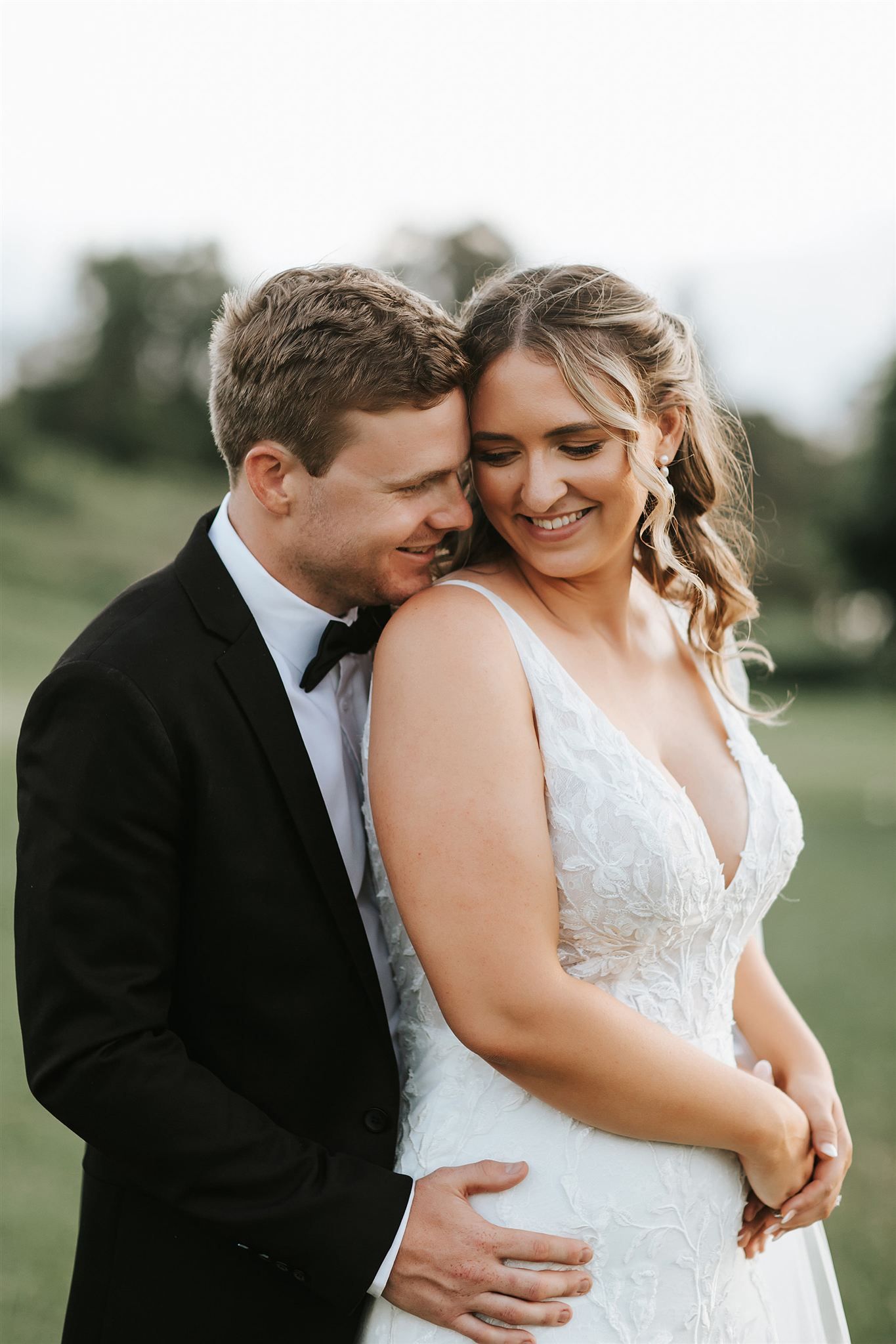 Groom in a black tuxedo embraces bride in a white beaded wedding dress, smiling. Green field backdrop in Northern NSW.