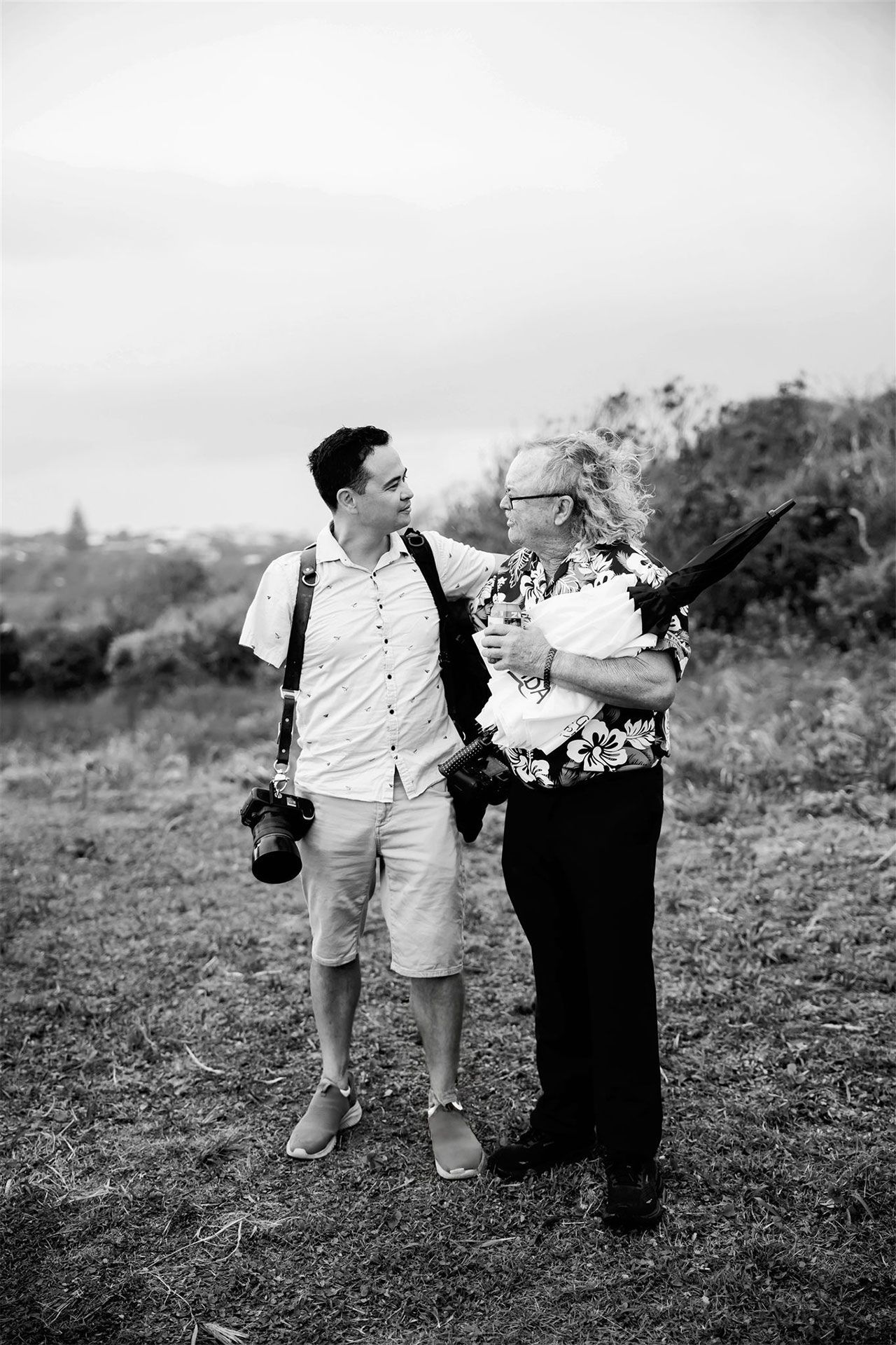 Denis Banks standing on a path in a wooded area, smiling. Wearing light-colored pants, a shirt, and carrying a camera.
