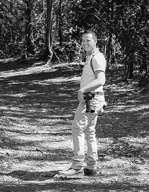 Denis Banks on a path in a forest, holding a camera, smiling. Black and white.