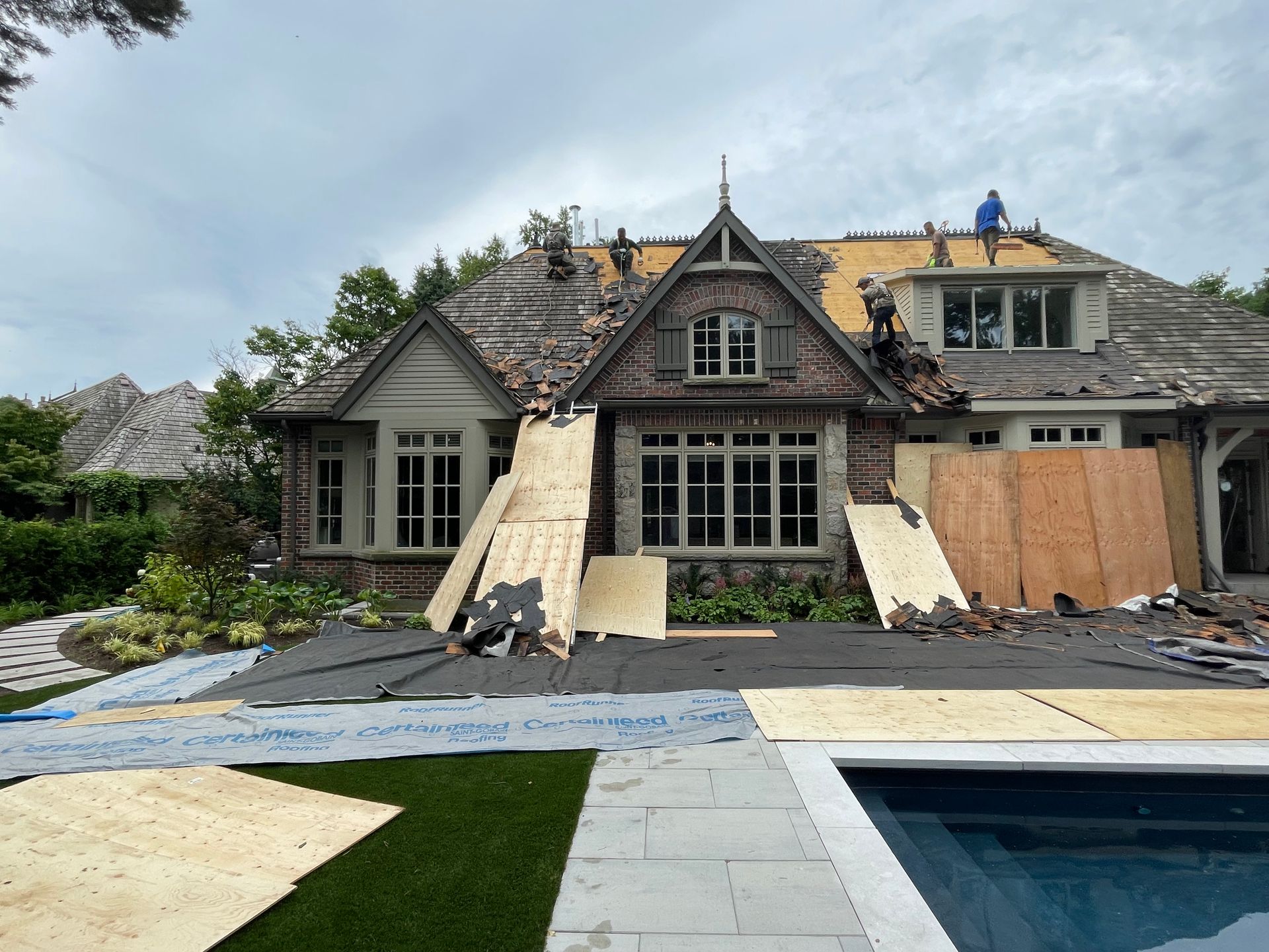Construction workers replace the roof on a brick house featuring a pool and green lawn in the backyard.