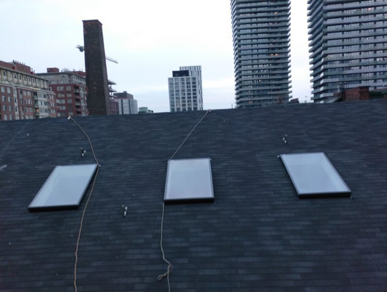 Three rectangular skylights on a dark shingled roof with city buildings in the background.