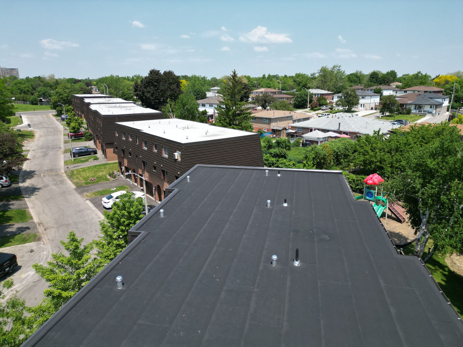 Overhead view of several buildings with black roofs. Houses in the distance, blue sky.