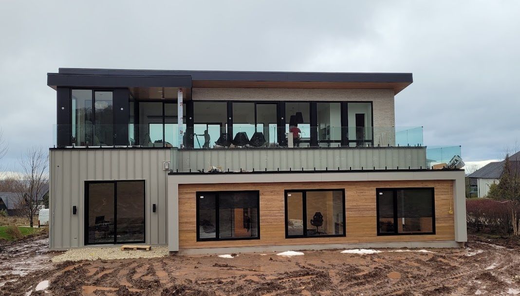 Modern two-story house with large windows, glass balconies, and wood accents, set in muddy, overcast landscape.