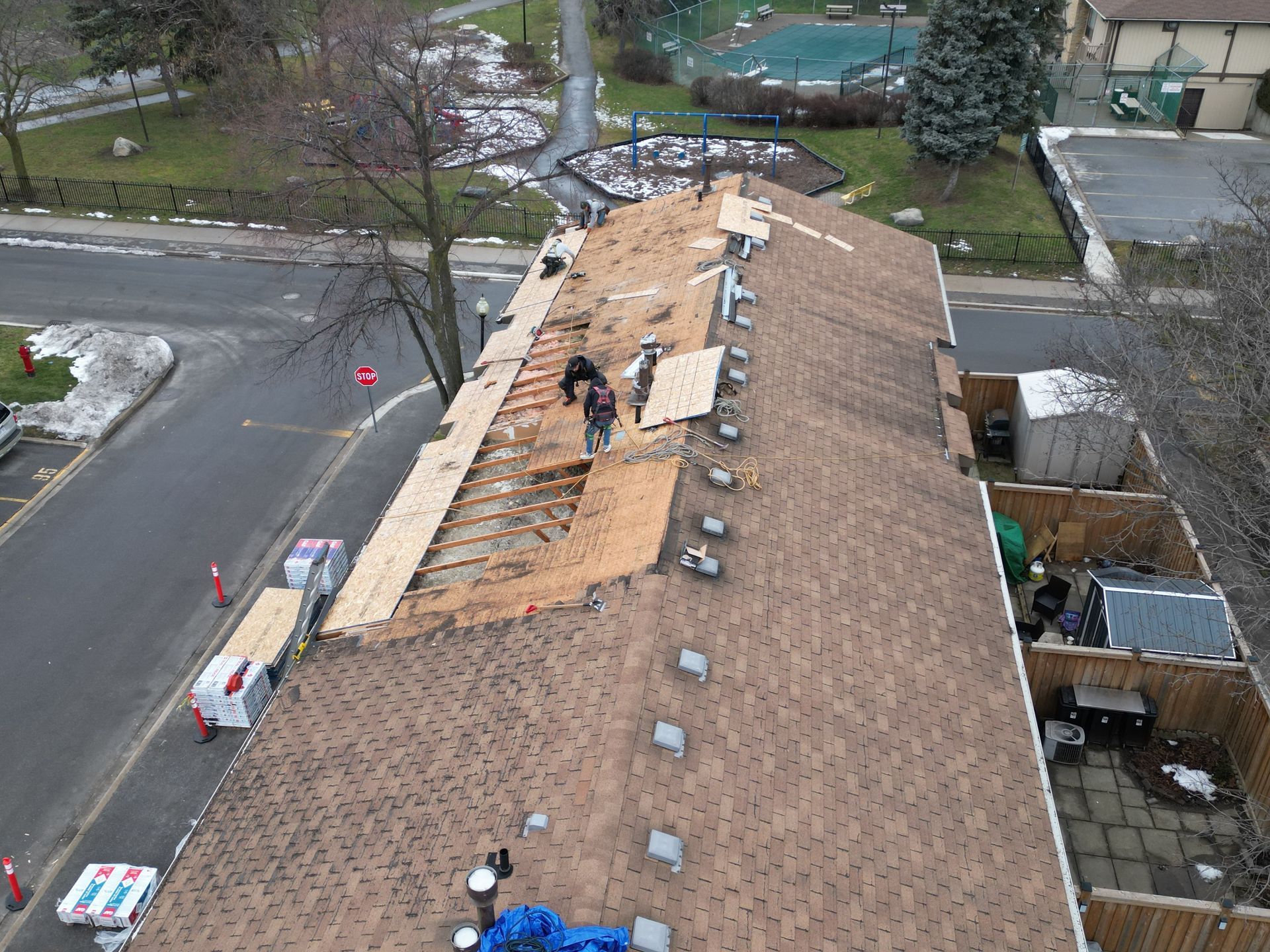 Roofers repair a section of a shingled roof on a multi-family building next to a street and parking area.