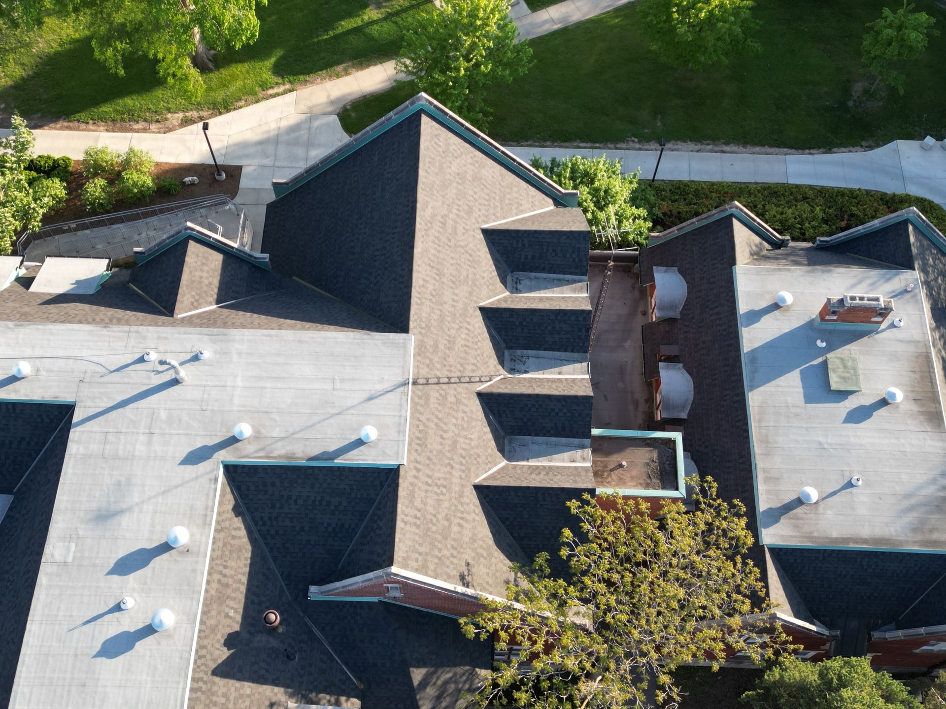 Overhead view of a building with asphalt shingles and flat roofs. Trees and a sidewalk are visible in the background.
