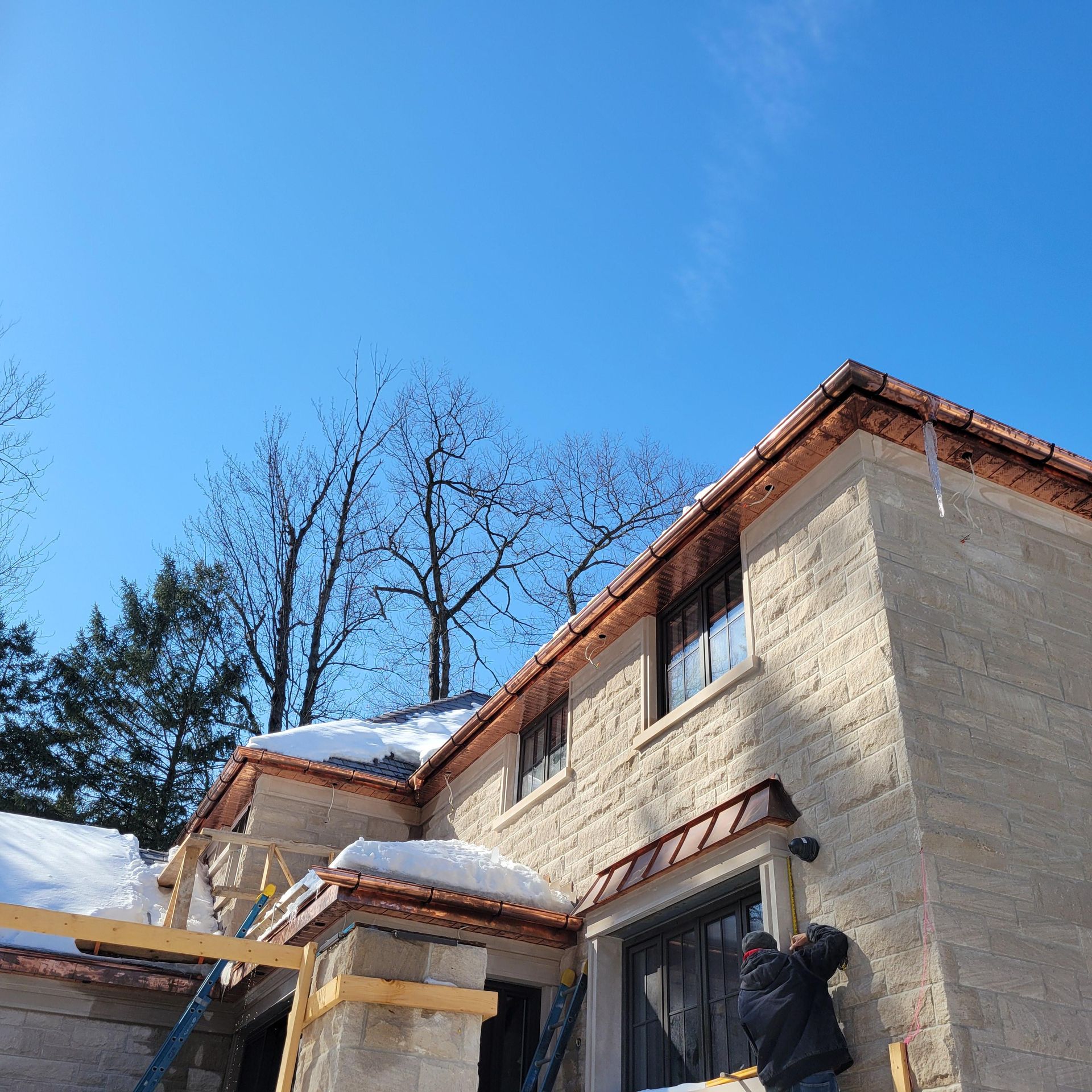Construction on a two-story tan stone building with copper gutters, a person working on the exterior under a blue sky.