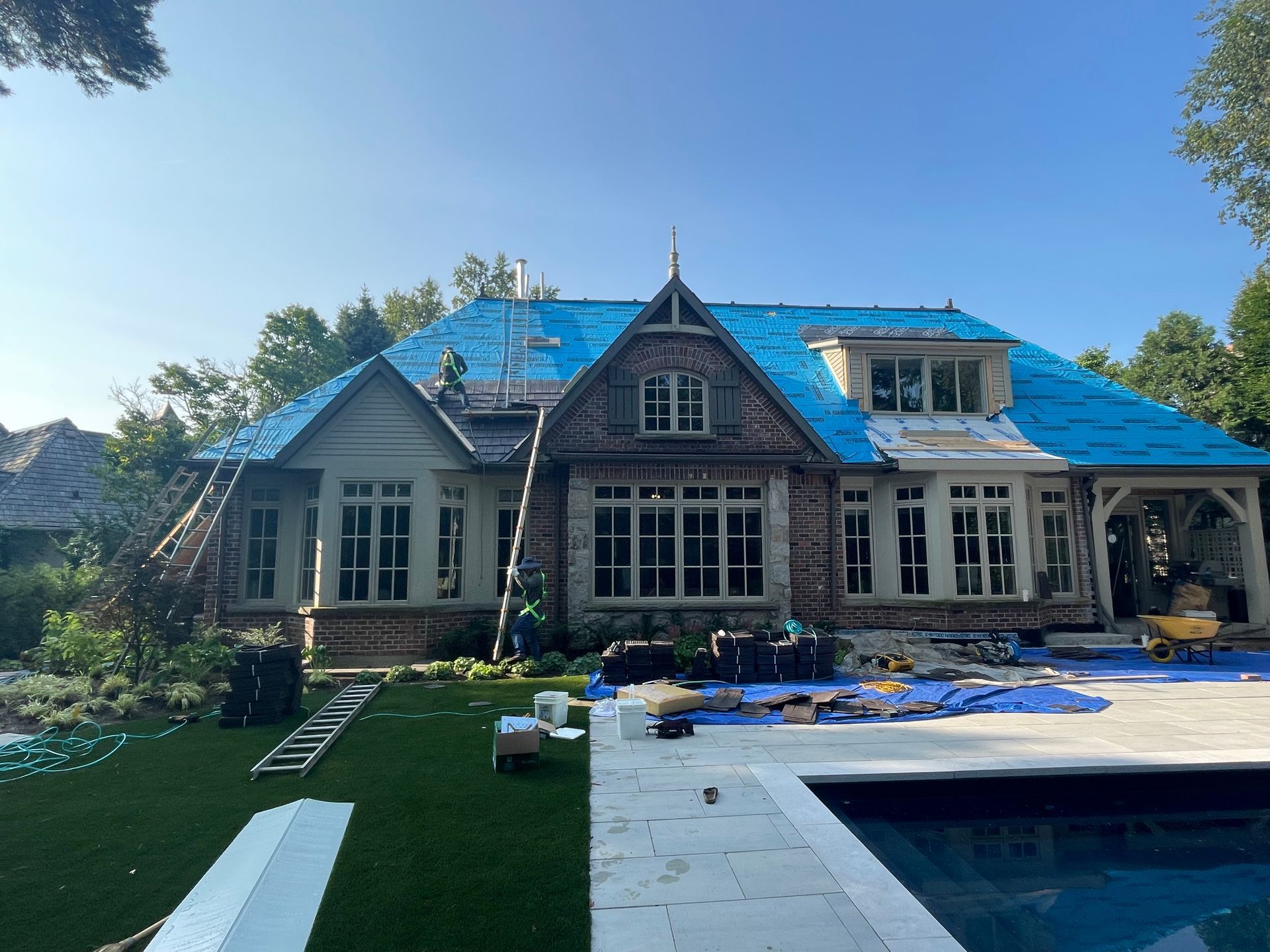 House under construction; blue tarp on roof, workers, pool, green lawn, sunny day.