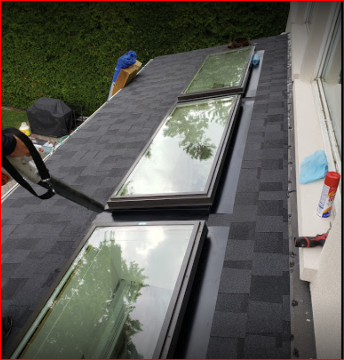 Three rectangular skylights installed in a row on a dark shingled roof next to a white building wall.