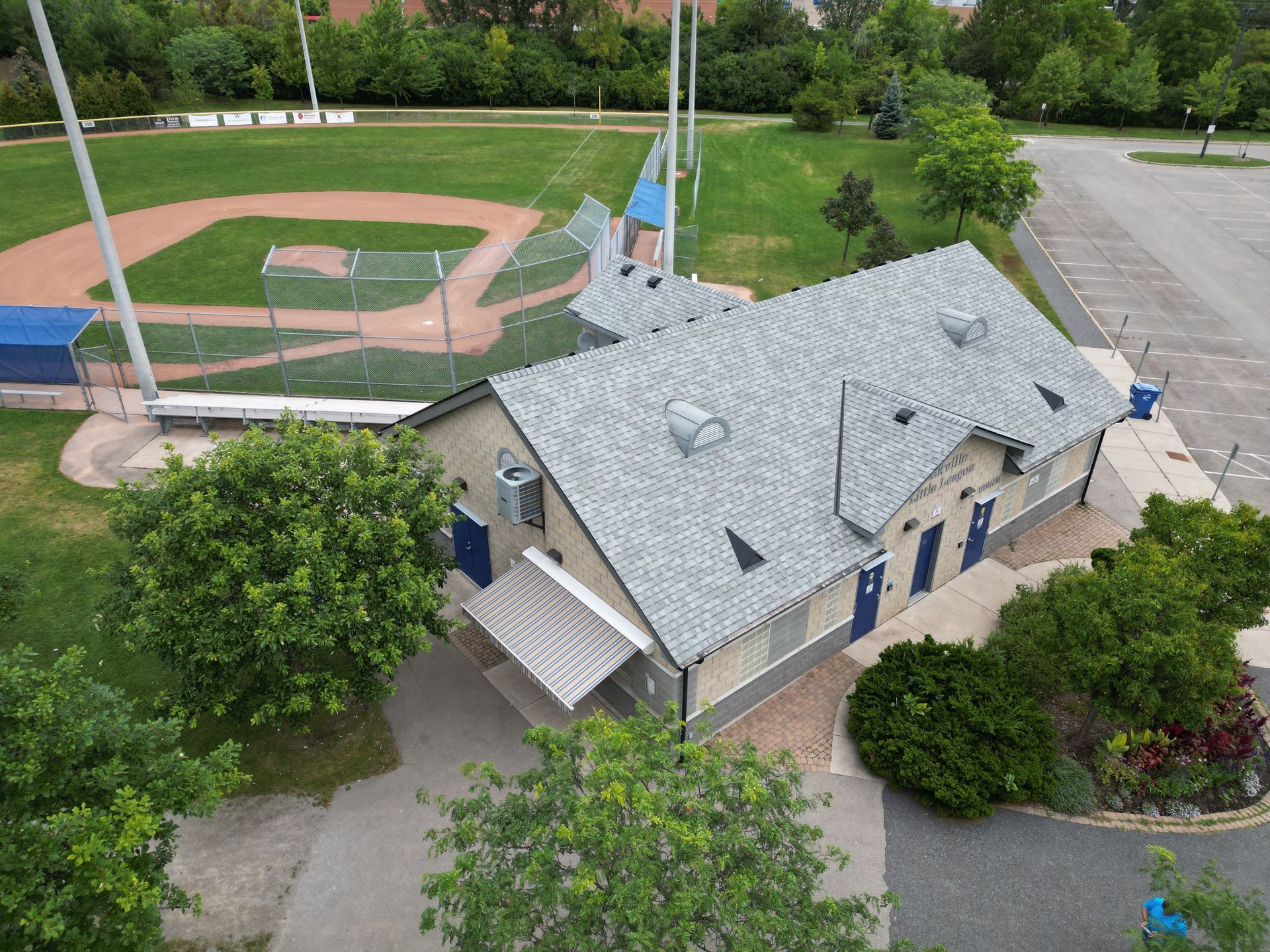 An aerial view of a baseball diamond next to a single-story stone building with a gray shingled roof and blue doors.