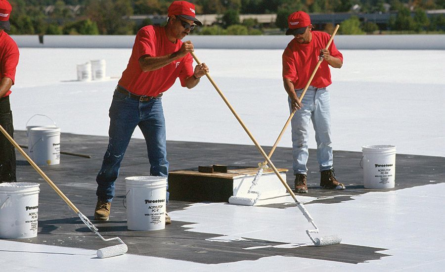 Workers applying white sealant to a flat roof with rollers. They wear red shirts and hats; buckets of sealant are nearby.