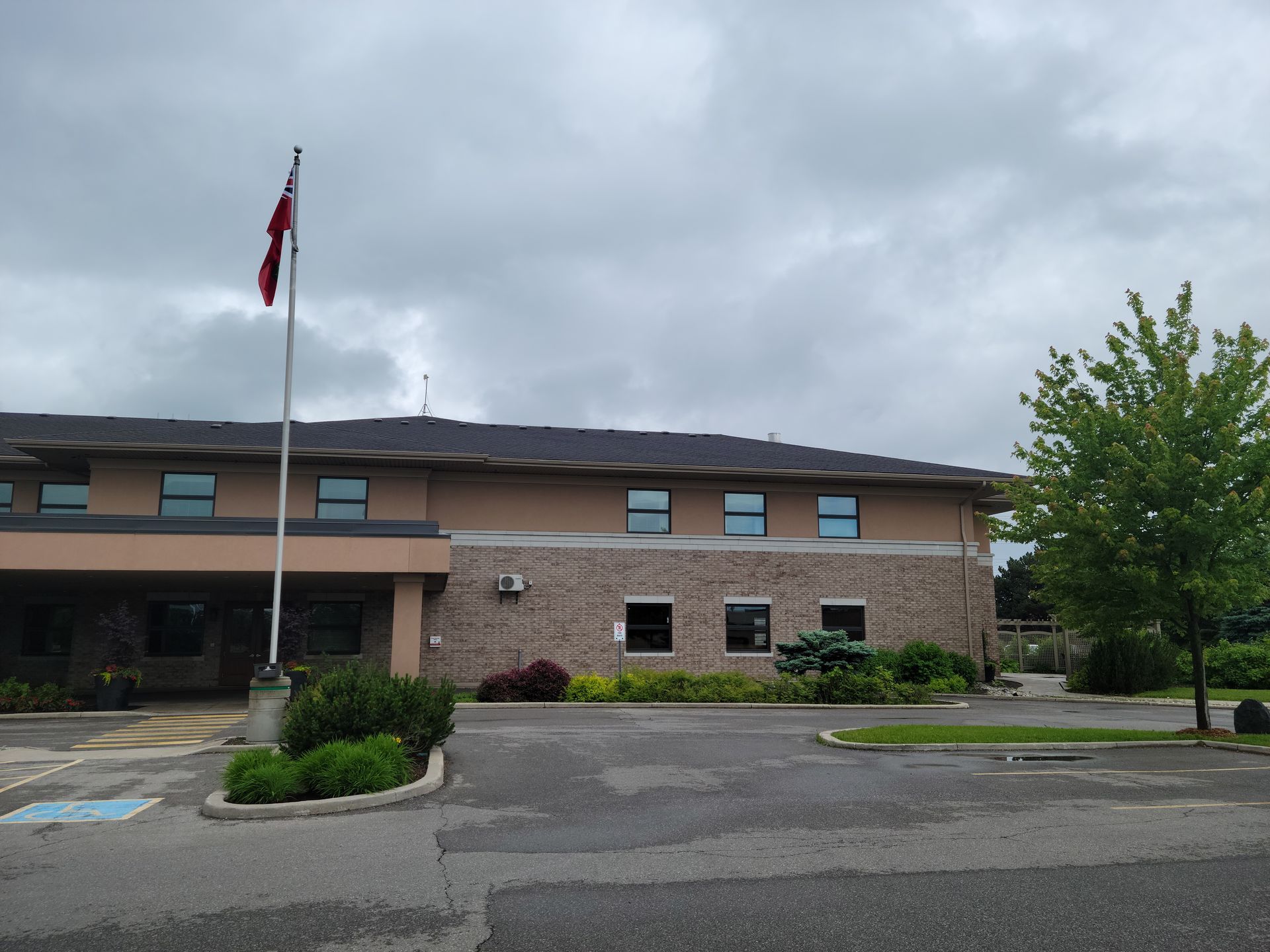 Two-story building with brick facade, Canadian flag, cloudy sky, and small tree in front.