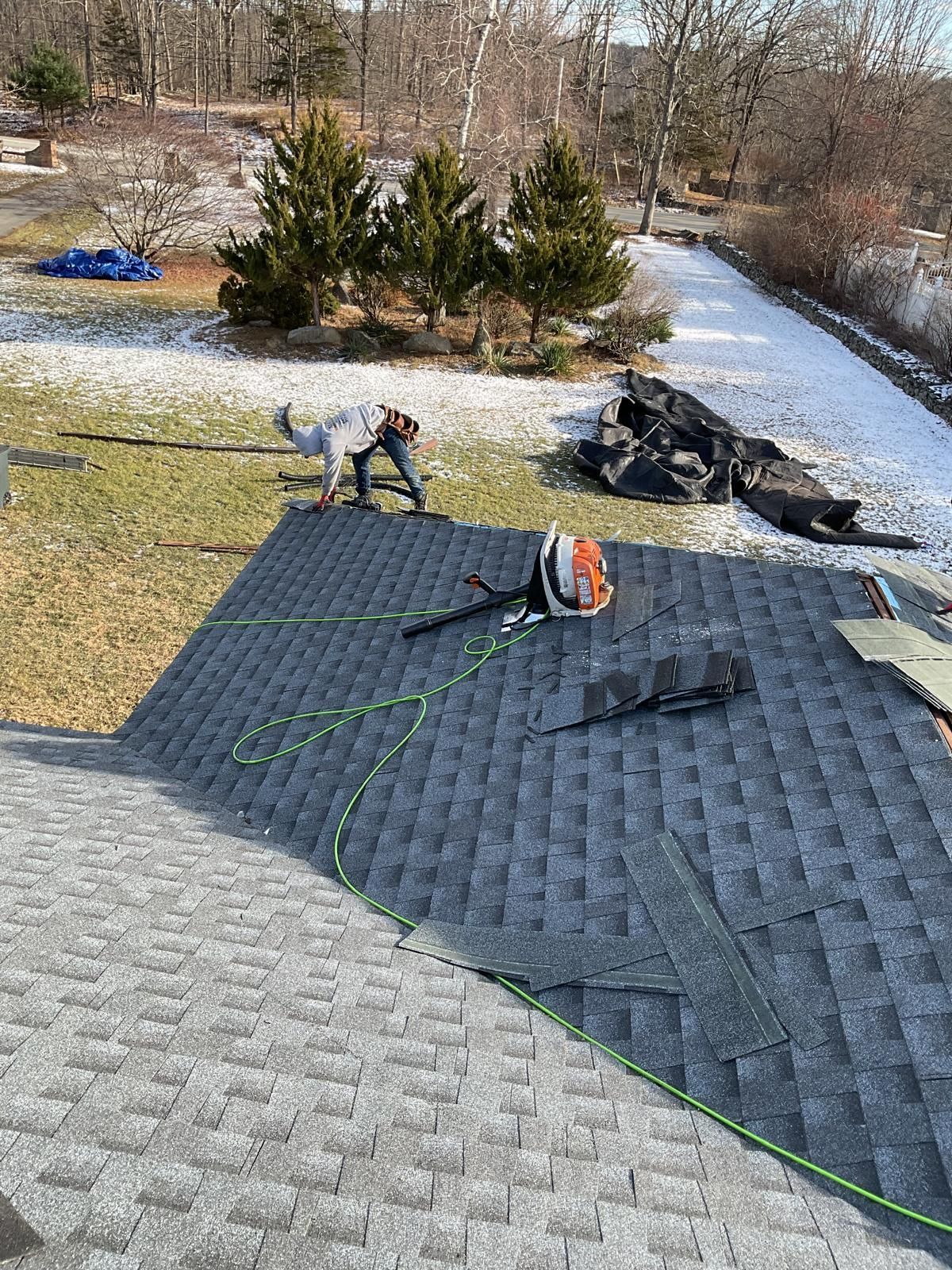 A worker repairs a gray shingled roof, while another person stands on the snowy ground nearby with construction debris.