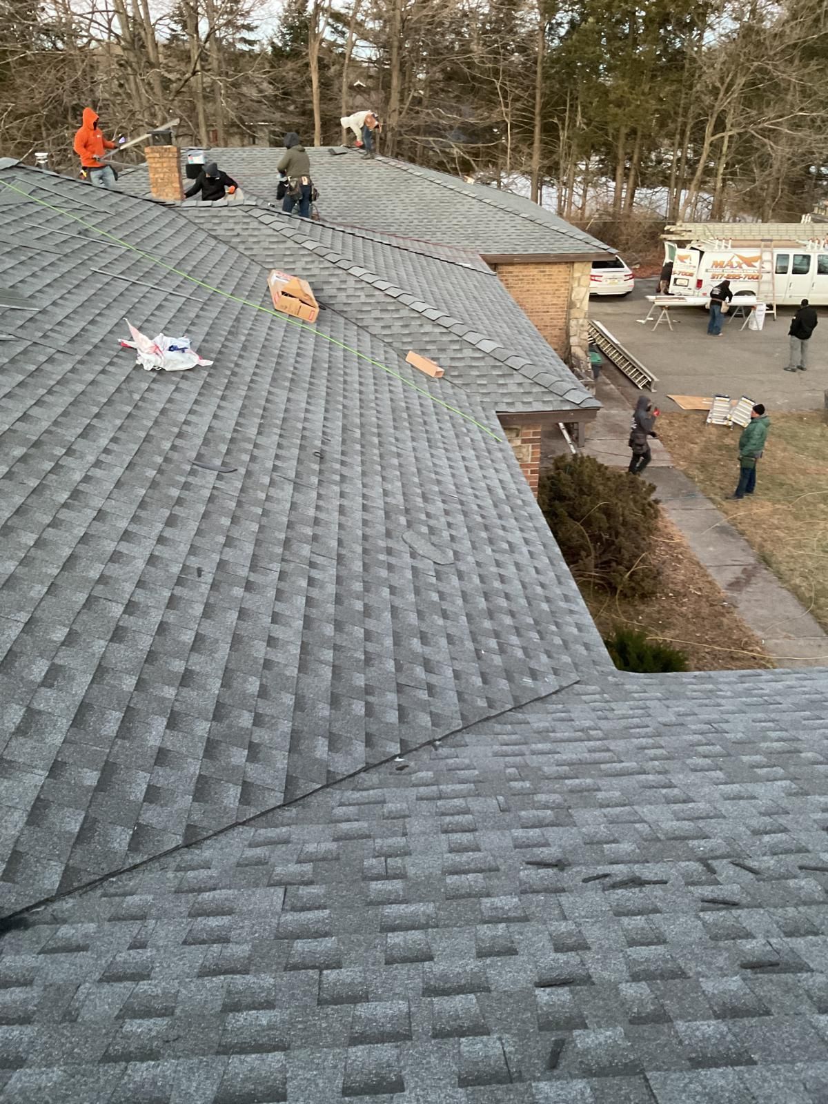 Several construction workers install shingles on a residential roof while others stand on the ground nearby.
