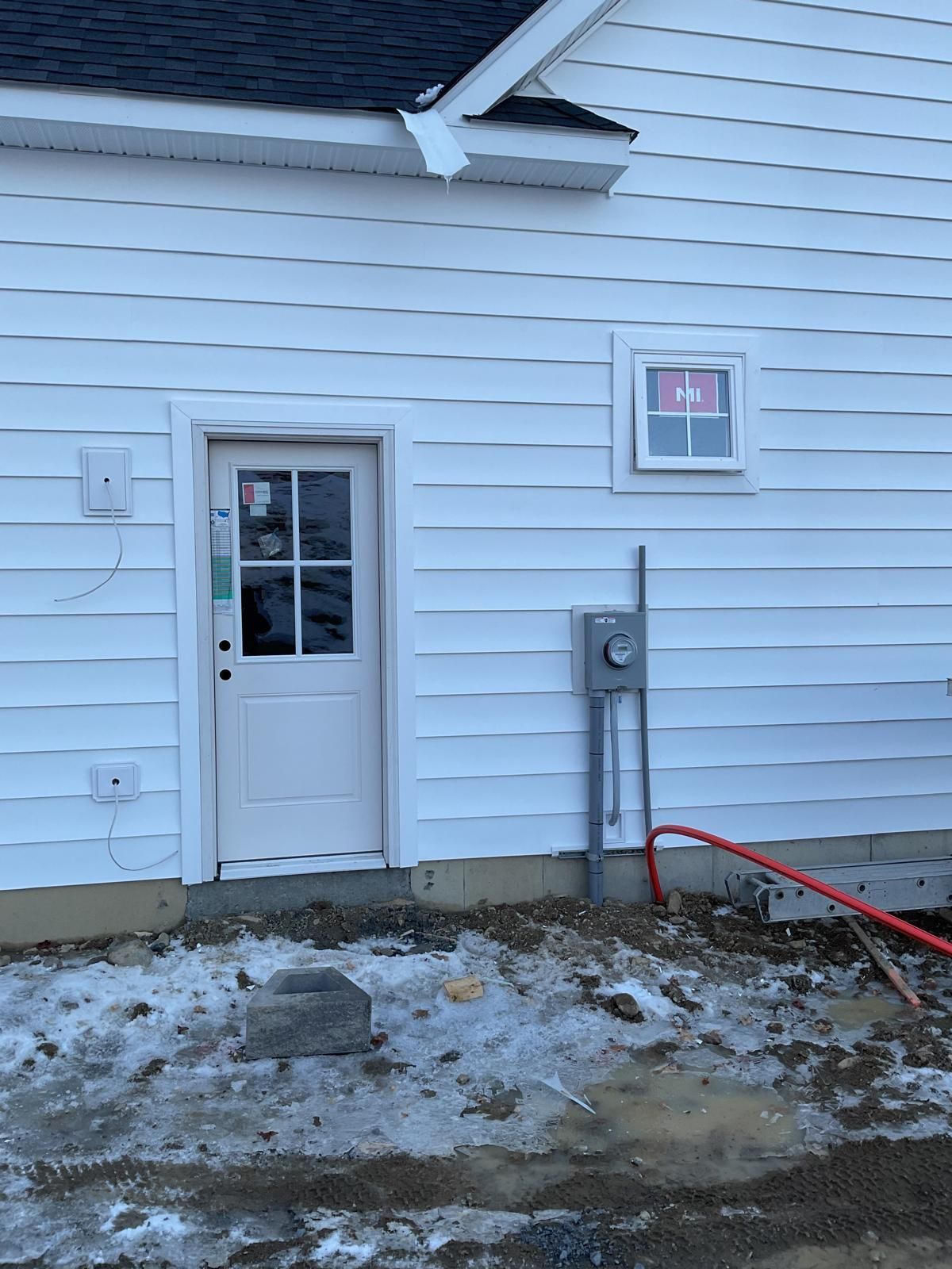 White siding house exterior with a door, a small window, and an electrical utility box above uneven, muddy, snowy ground.