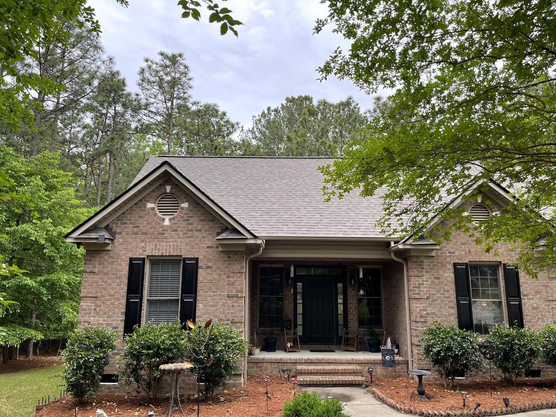 A brick house with a gray roof and black shutters is surrounded by trees.