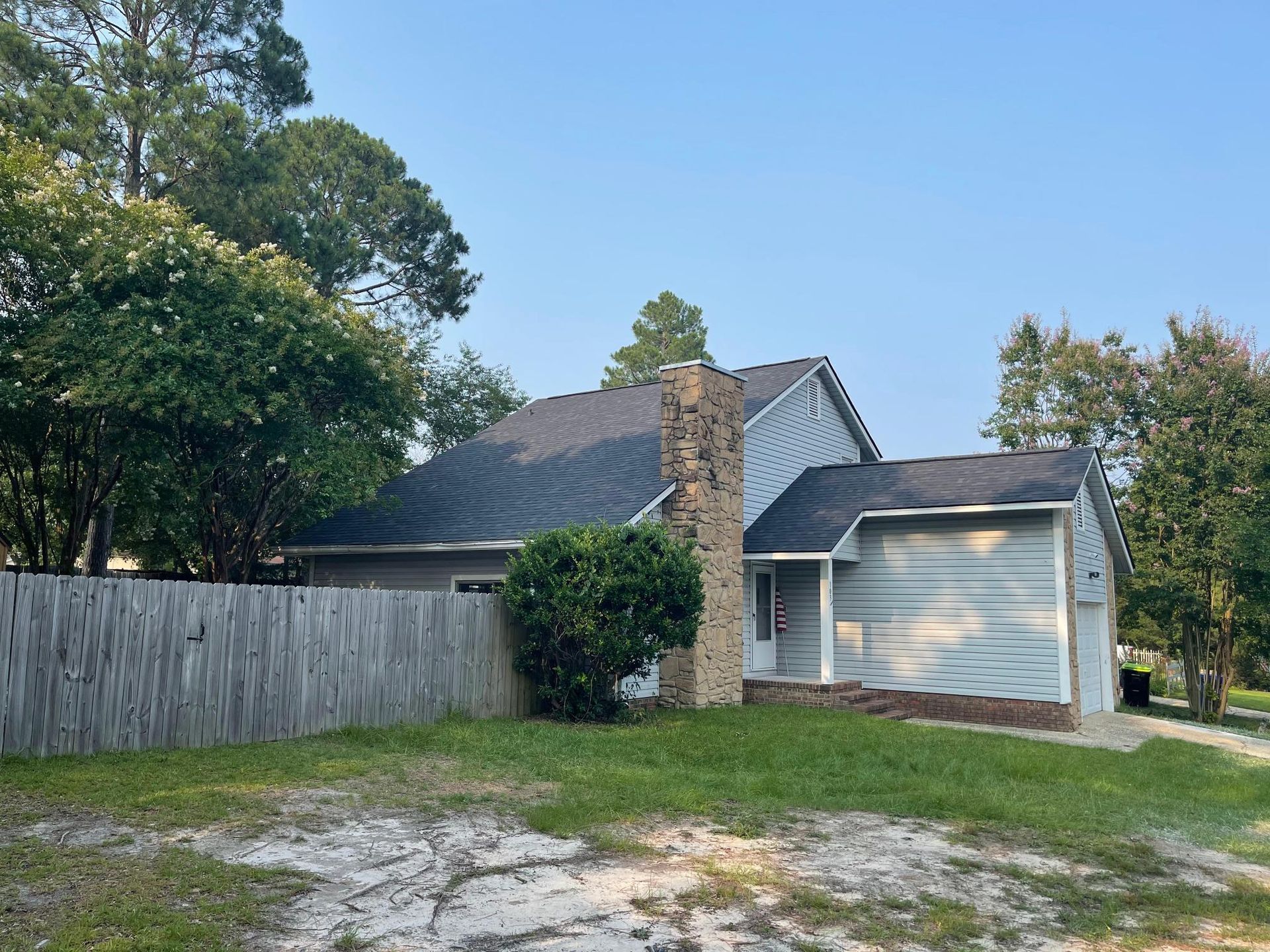 A white house with a black roof is surrounded by trees and a wooden fence.