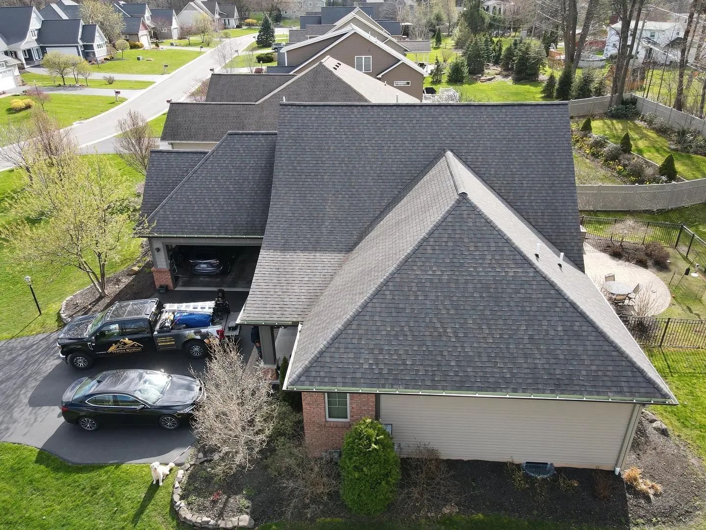 Aerial view of a suburban house with dark shingle roof, driveway, and parked cars.