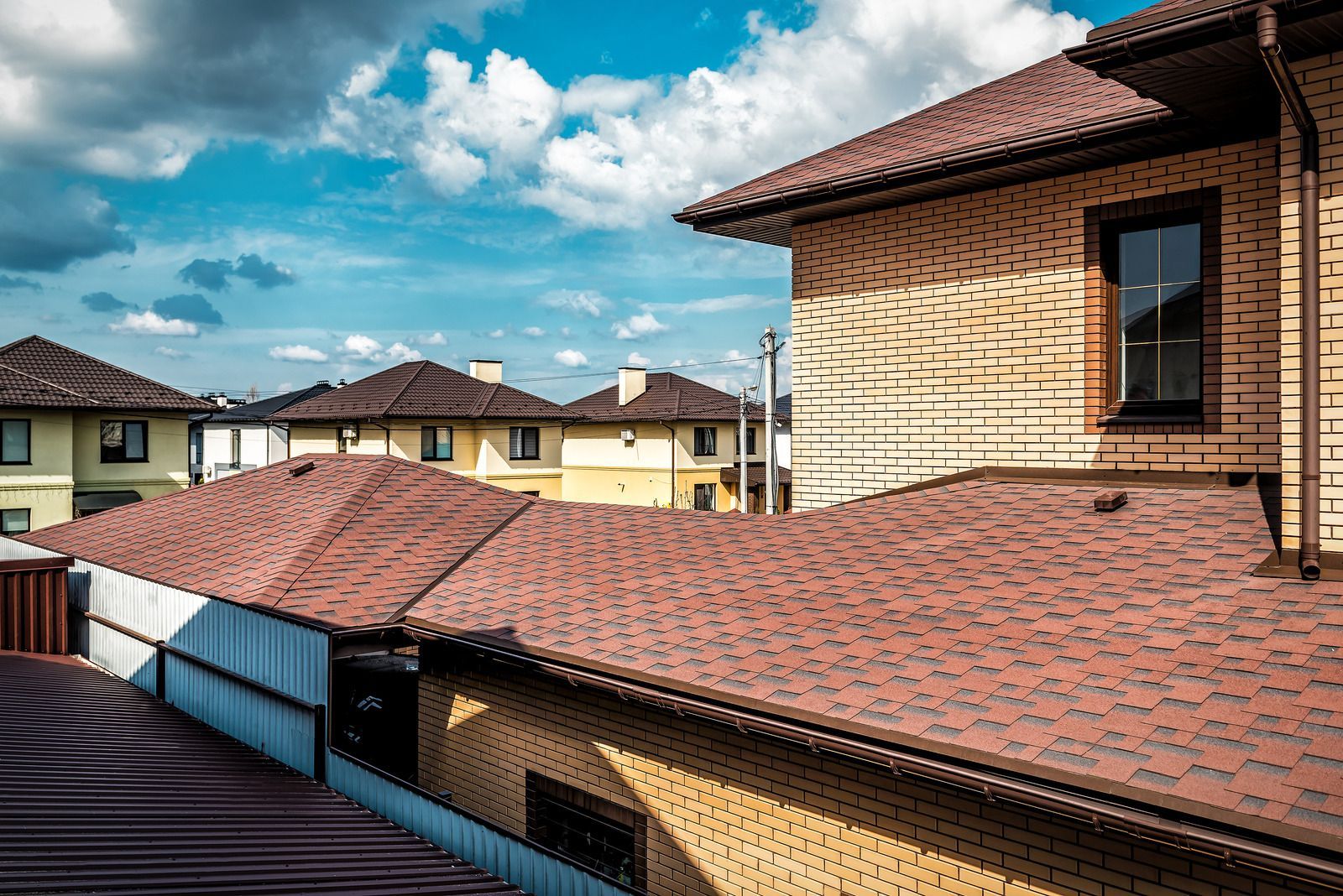 Low-angle view of modern houses with brown shingled roofs and light-colored brick walls under a blue sky with clouds.