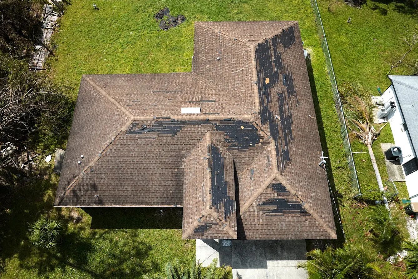 Aerial view of a house with a brown roof and visible damage, surrounded by green grass.
