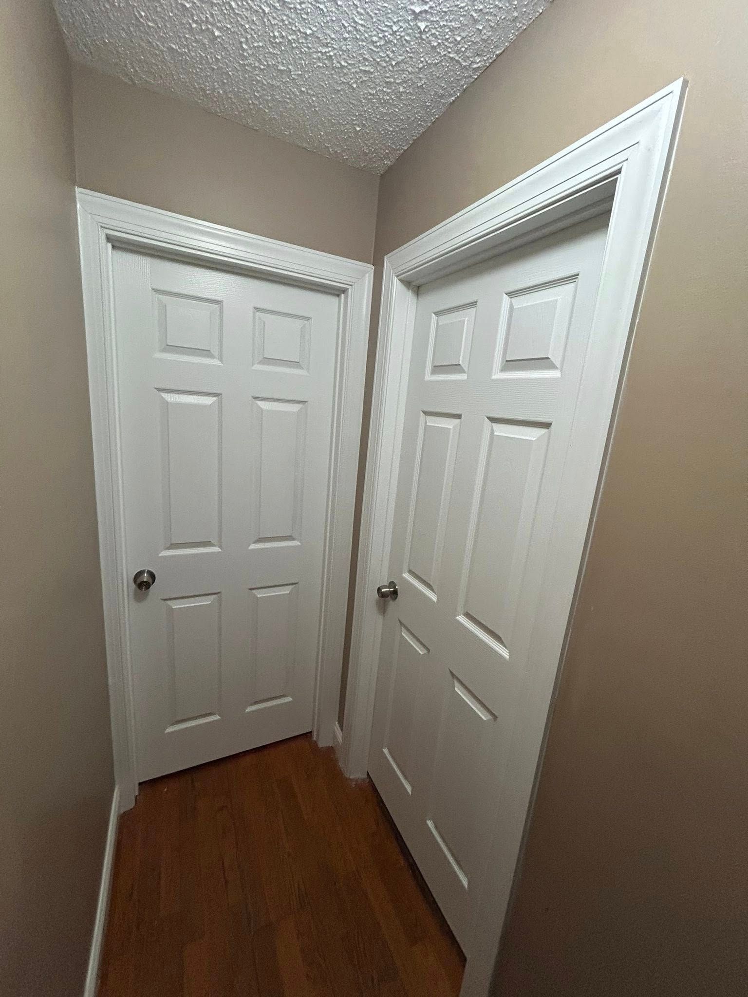 Two white doors in a narrow hallway with hardwood floors and tan walls.